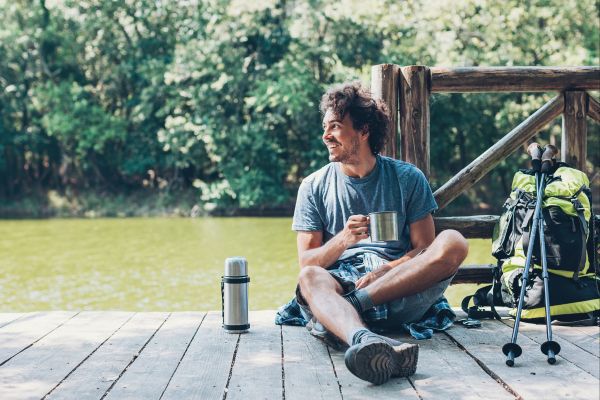 Un homme assis au bord d'un lac, profitant de la nature avec une tasse en métal et un sac à dos, illustrant un mode de vie zéro déchet et écoresponsable.