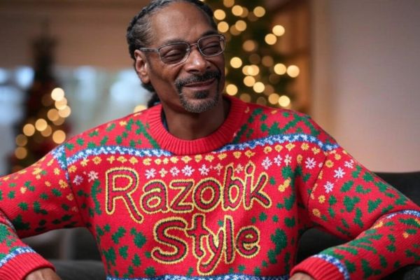 Snoop Dogg portant un pull moche Razobik rouge avec l’inscription "Razobik Style" devant des décorations de Noël.